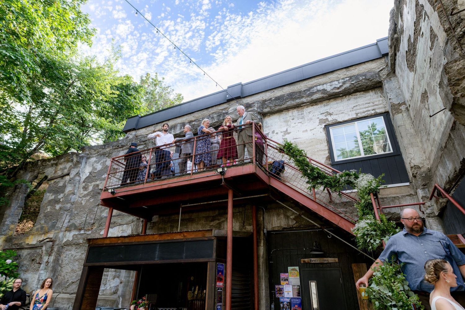 Guests gather on rustic stone building balcony during outdoor wedding reception celebration.