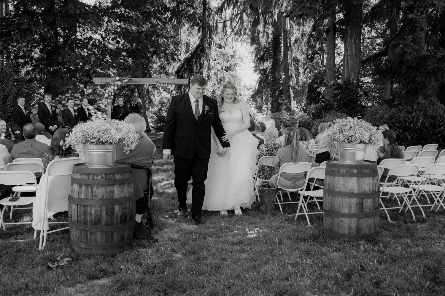 Black and white photo of bride and groom walking down the aisle at an outdoor rustic barrel-decorated wedding.