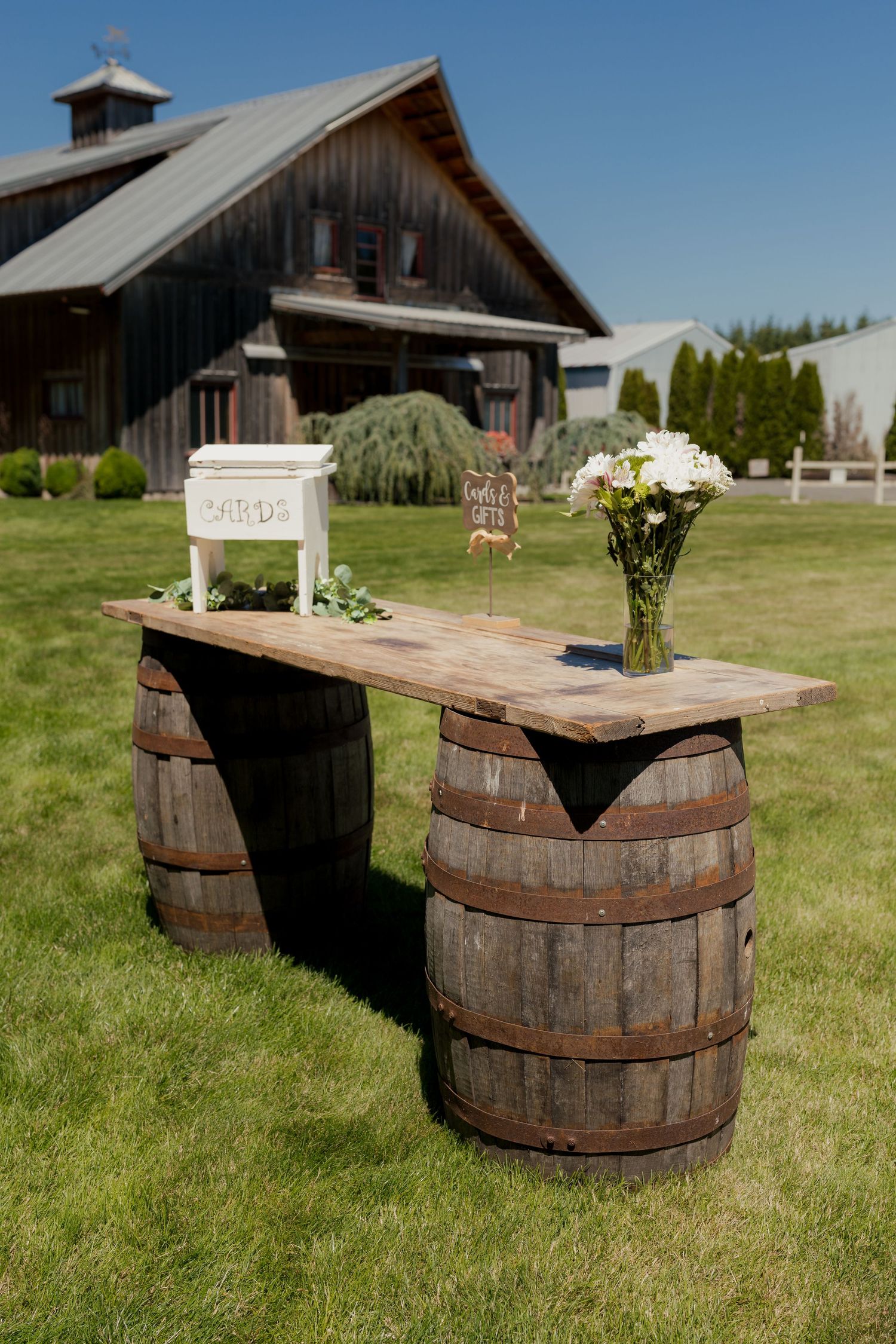 Rustic wedding card table made from wooden barrels and a plank, decorated with flowers, set on a green lawn in front of a barn.