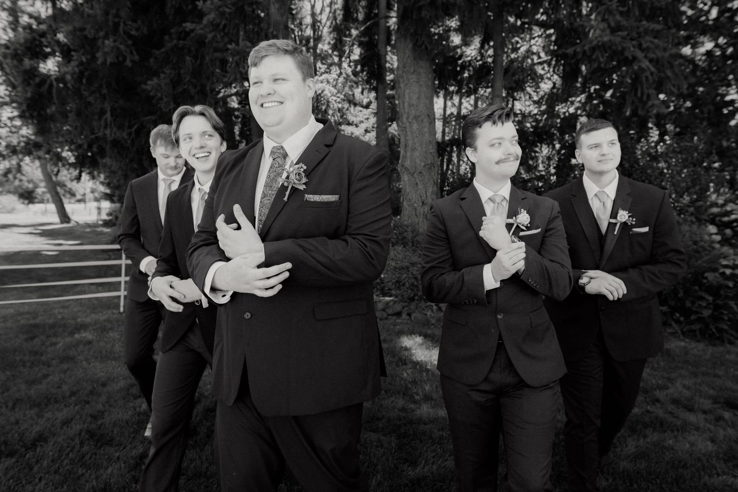 Black and white photo of groomsmen in dark suits walking together and adjusting cufflinks outdoors.