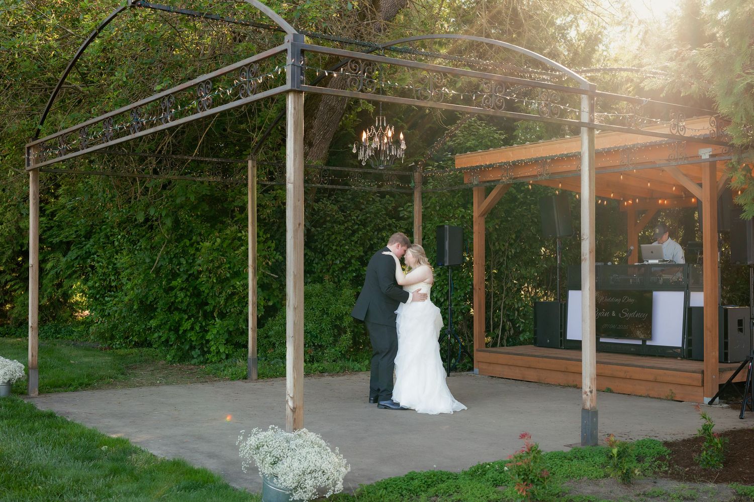 Bride and groom share romantic kiss under elegant chandelier pergola at garden wedding.