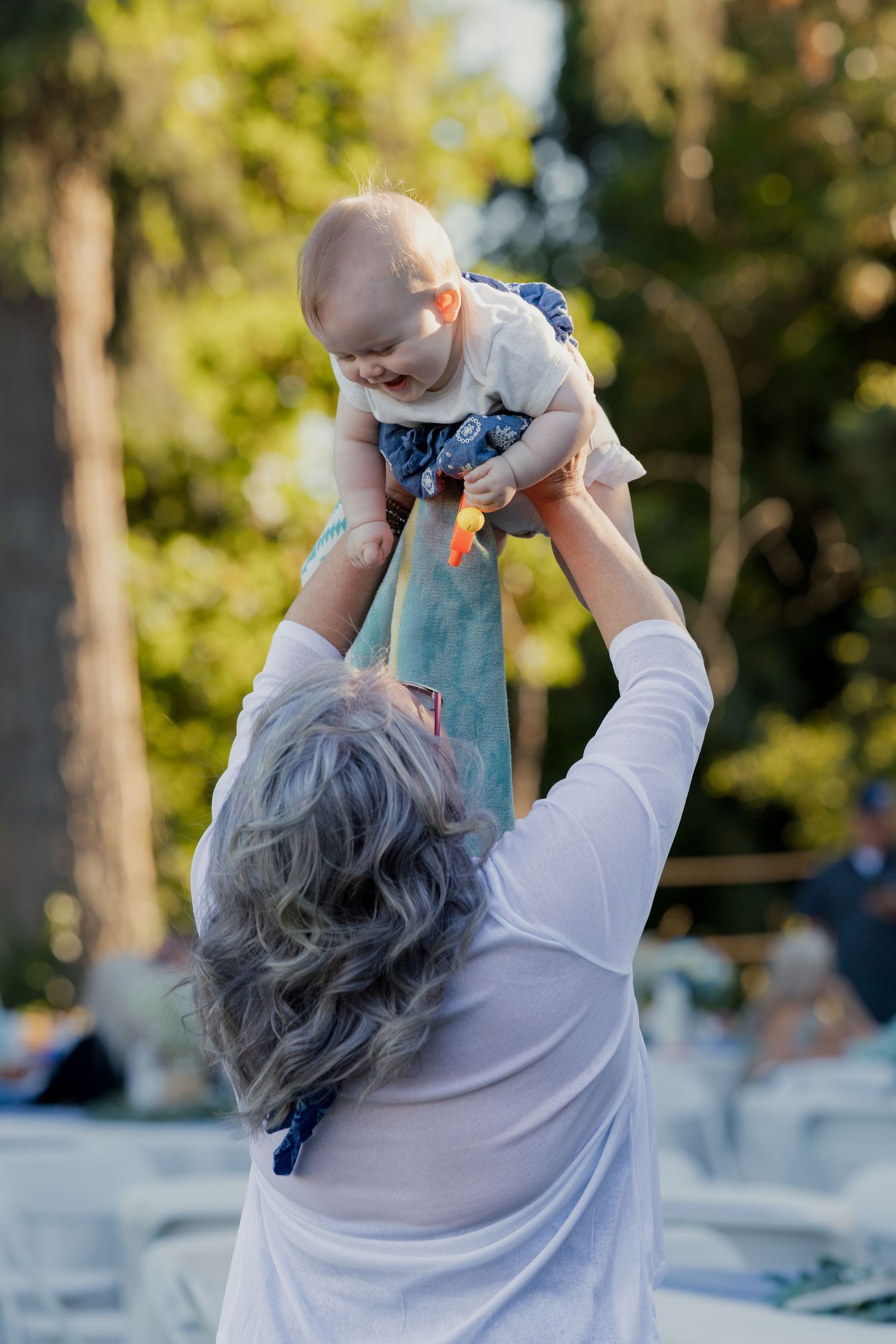 Grandmother joyfully lifts laughing baby into the air at an outdoor wedding reception.