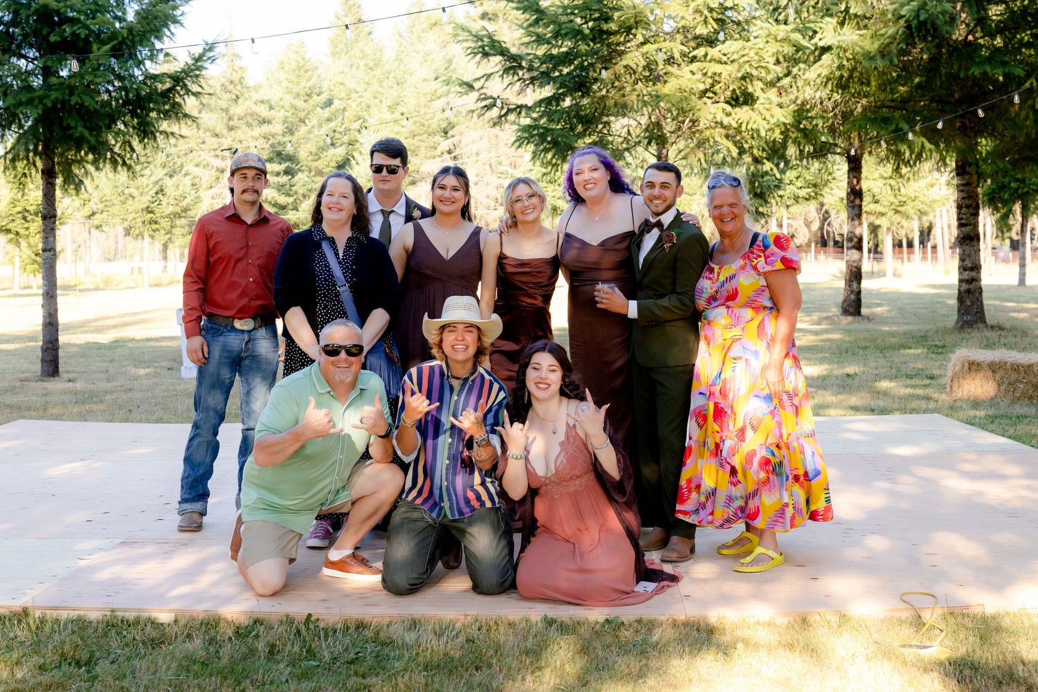Group of wedding guests posing together for a fun photo in a tree-lined outdoor venue on a sunny day.
