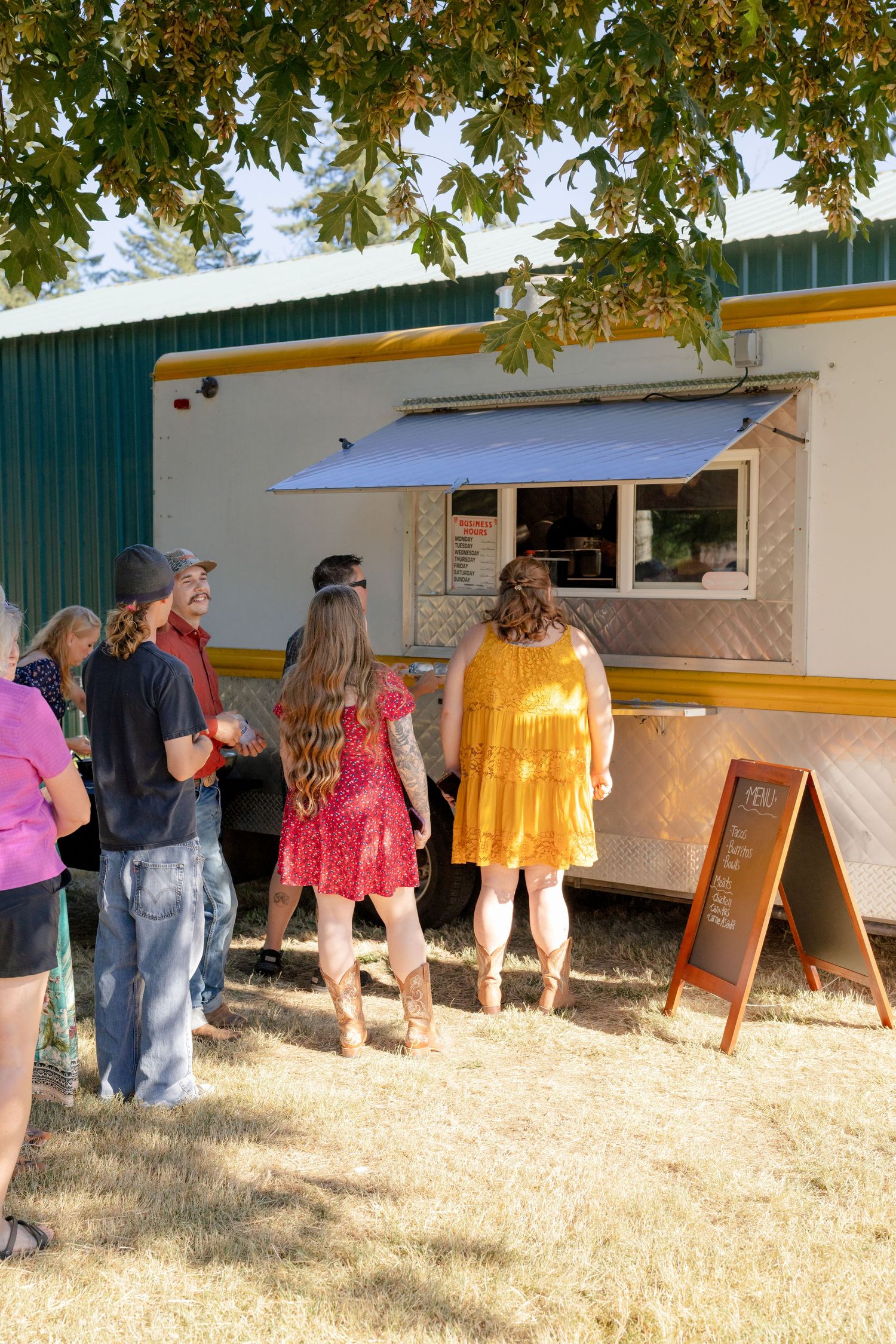 Wedding guests queue at food truck under leafy tree at sunny outdoor rustic wedding reception.