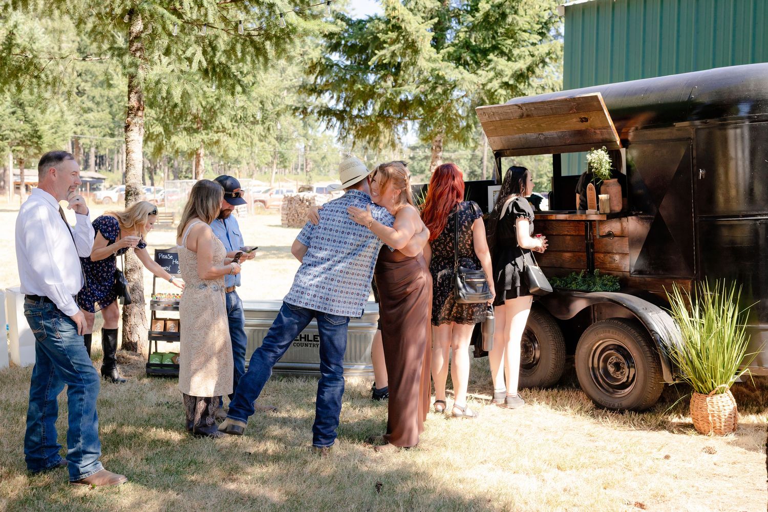 Wedding guests socializing near a vintage rustic bar truck outdoors surrounded by tall pine trees on a sunny day.