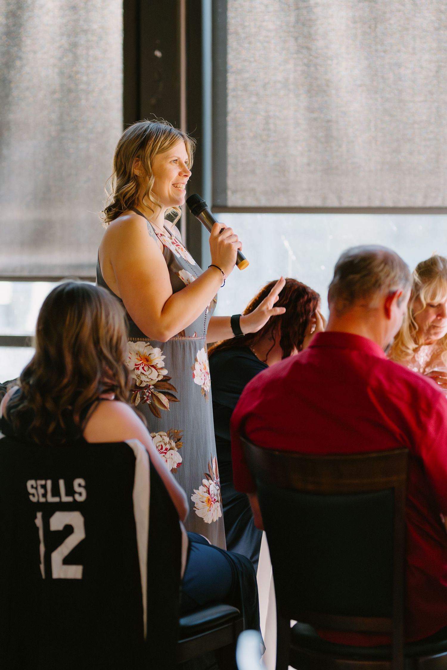 Woman in floral dress speaking into microphone at an indoor event with seated guests.