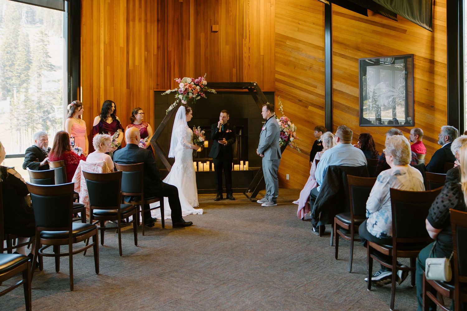 Indoor wedding ceremony with guests seated in wooden chairs, couple at altar with officiant, mountain views through windows.
