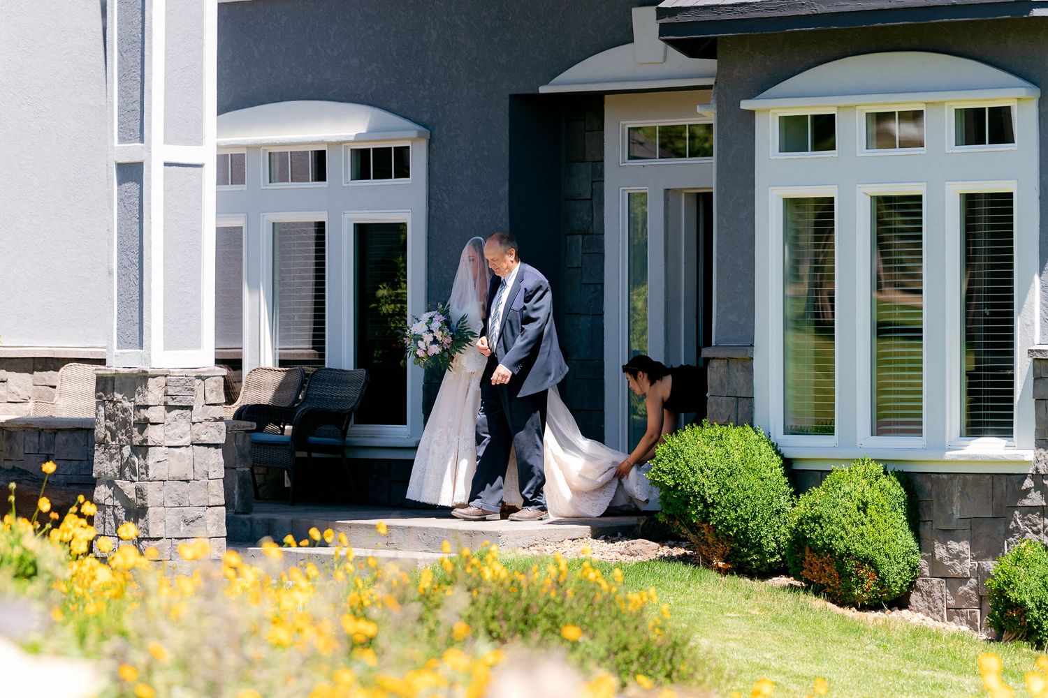 Bride and groom walk outside a gray stone house as an attendant helps carry the wedding dress train.