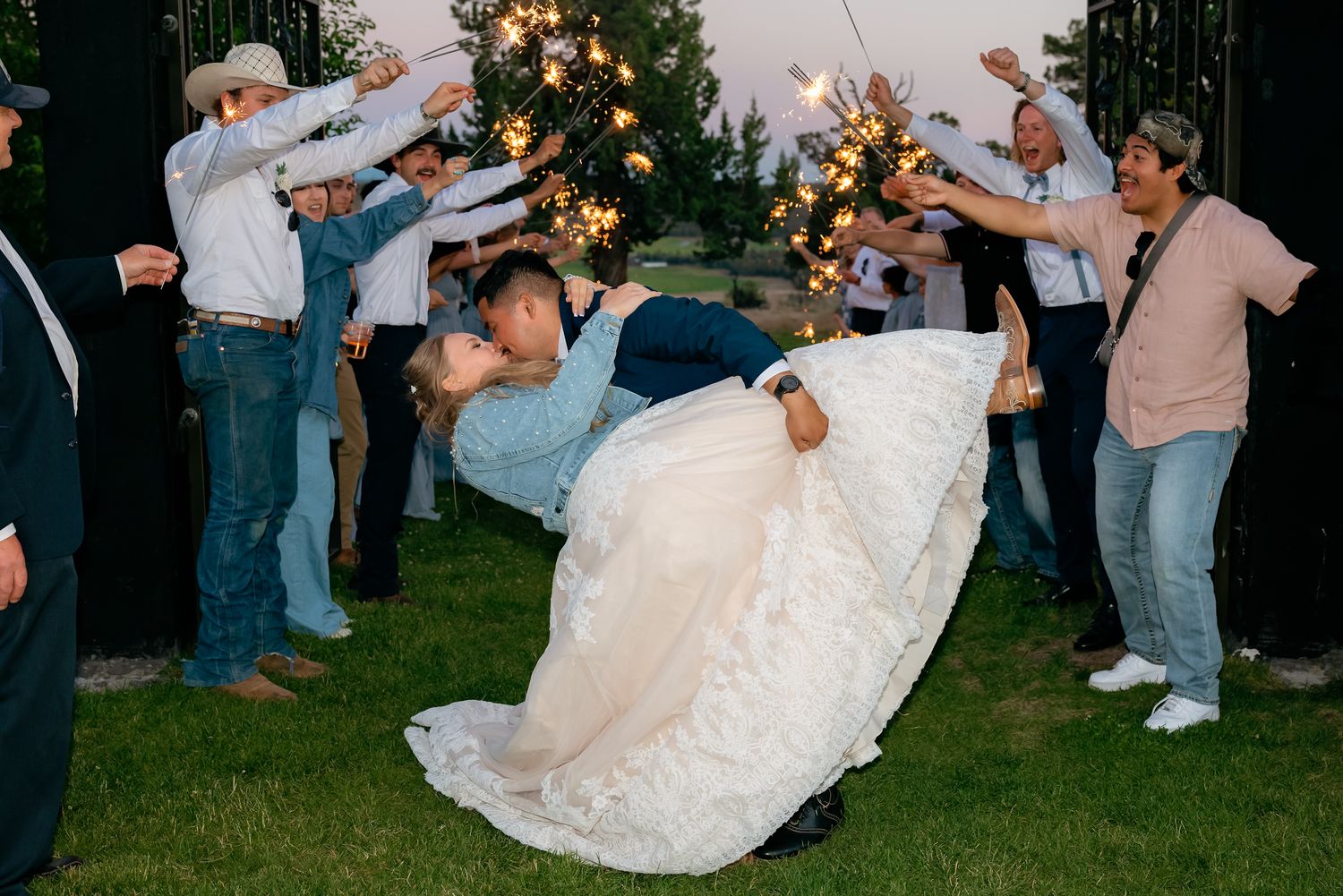 Bride and groom share a romantic dip kiss surrounded by guests holding sparklers at sunset.
