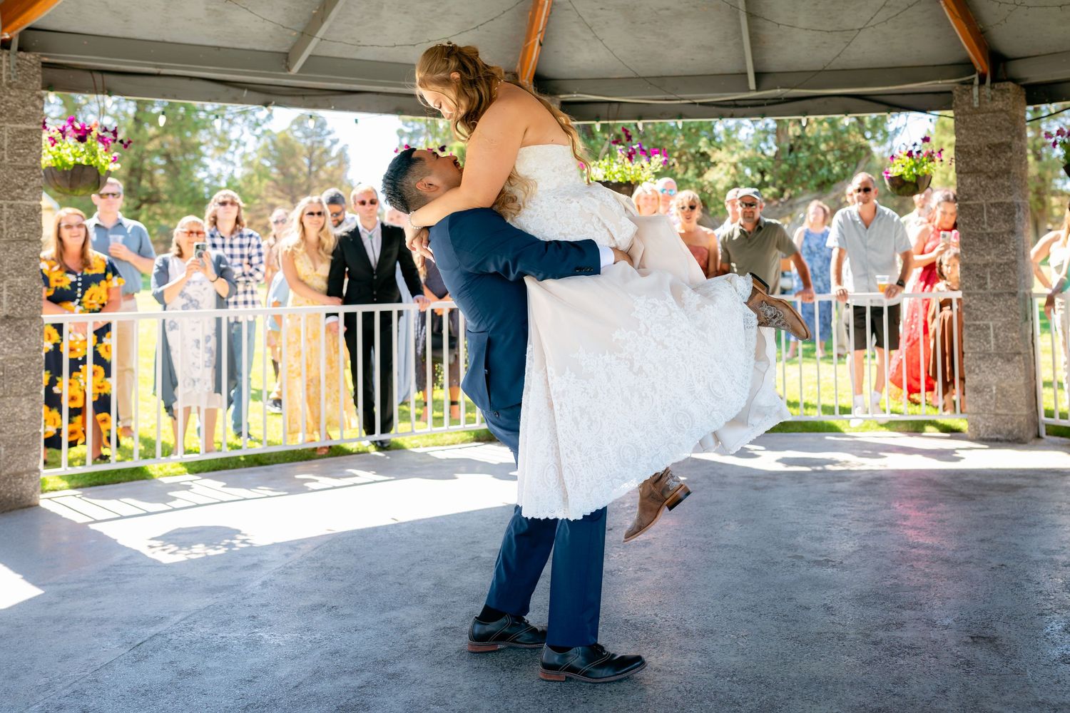 Groom lifts bride in white lace dress during first dance under outdoor pavilion.