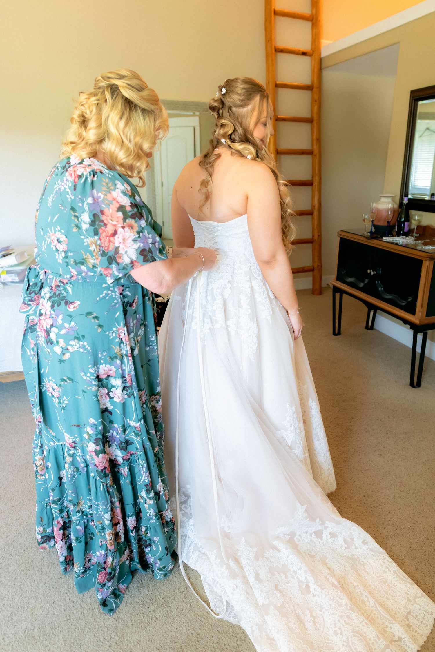 A woman in a floral dress helps a bride in a white lace gown get ready indoors before her wedding ceremony.