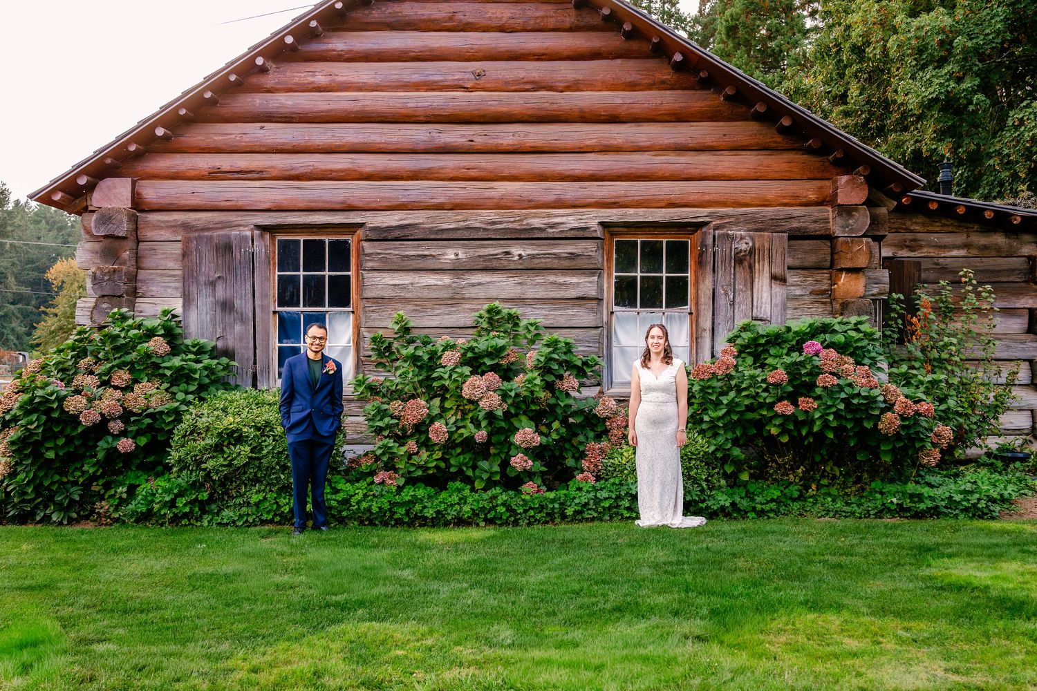 A rustic log cabin with red trim and climbing roses sits nestled among lush green gardens and trees.