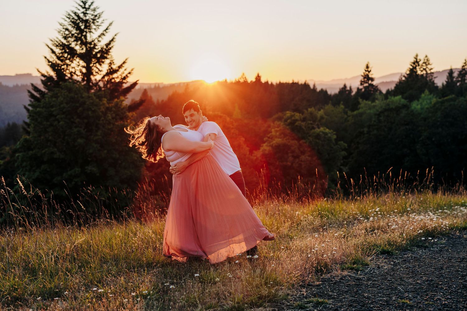A couple poses together in a field at sunset with evergreen trees in the background.