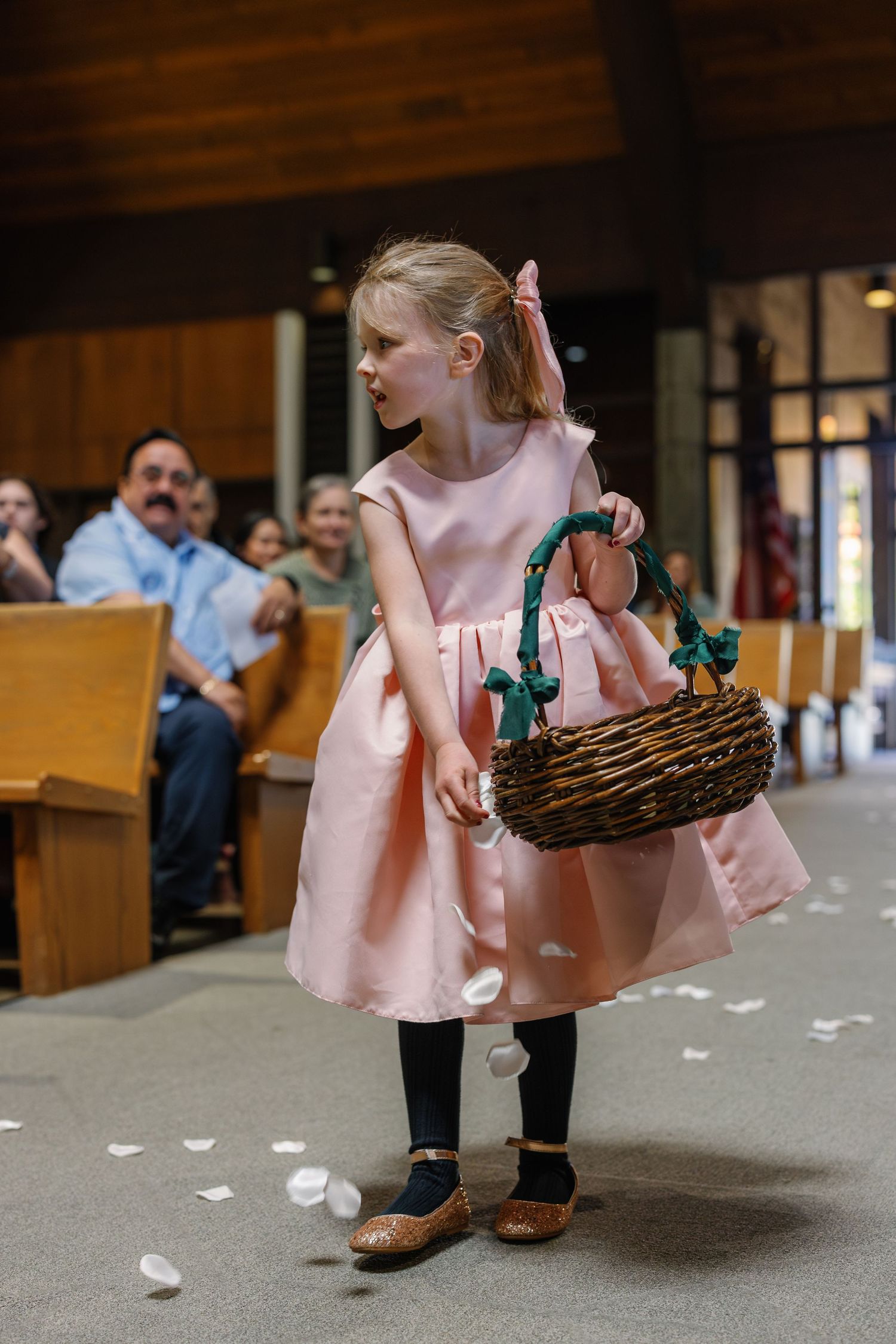 A flower girl in a pink dress carries a woven basket down the aisle while white petals scatter on the floor.