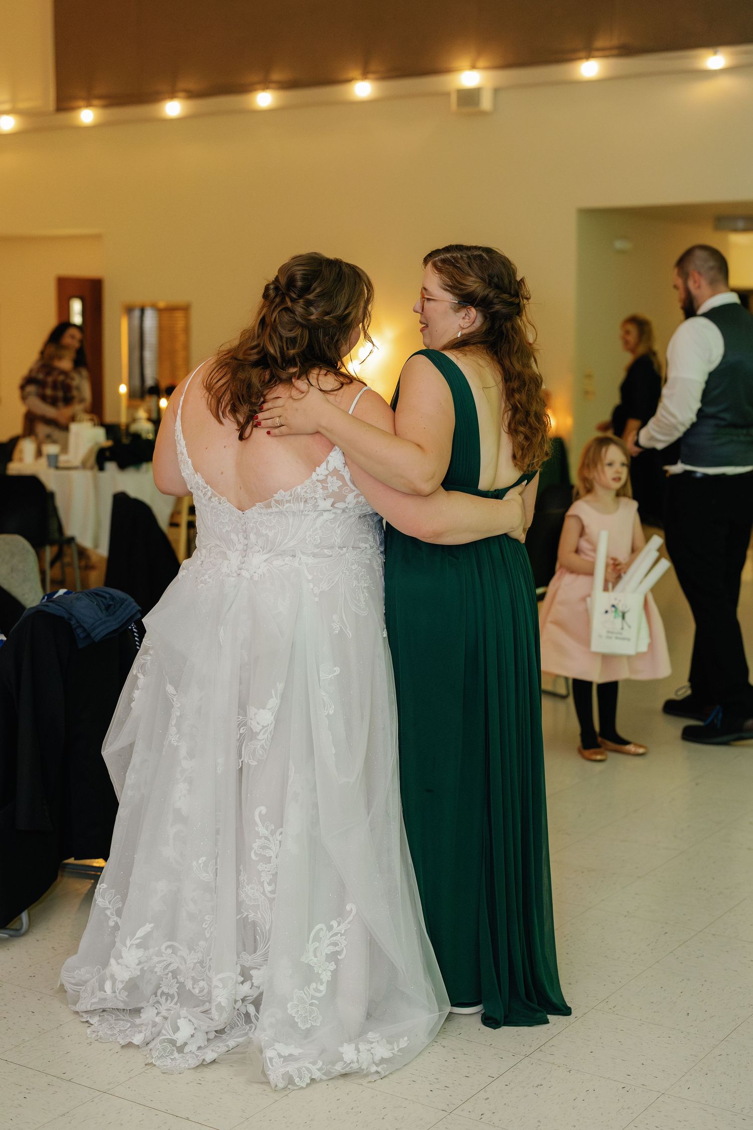 Two women embrace on the dance floor, one wearing a white wedding gown and the other in an emerald green dress.