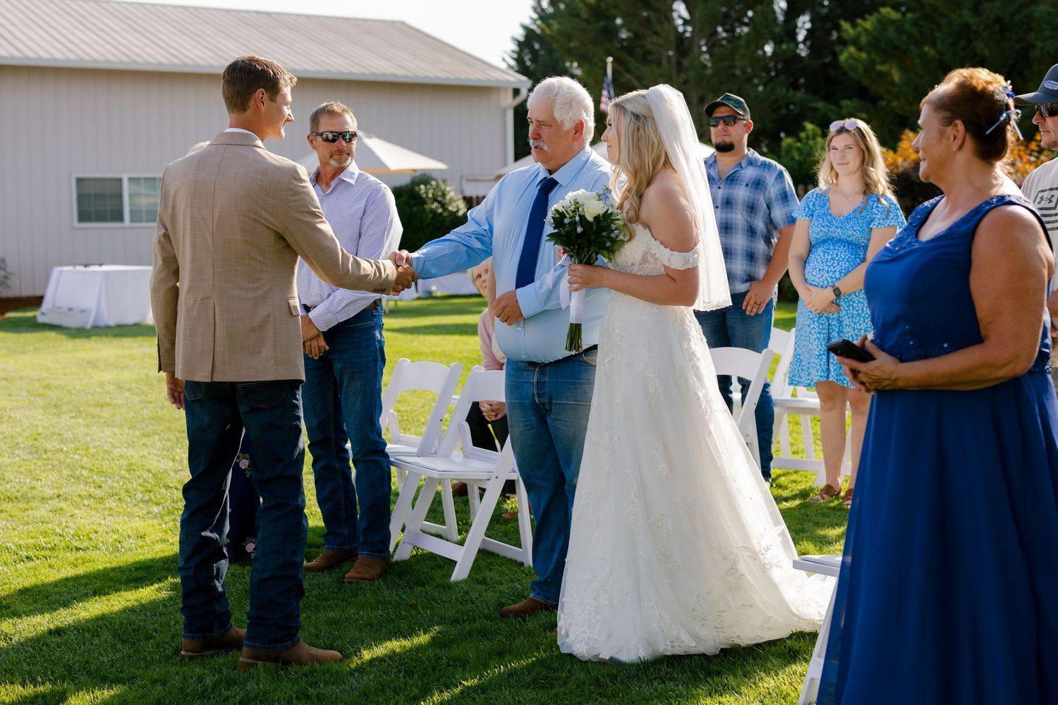Wedding guests mingling outdoors in blue dresses and casual attire near white chairs.