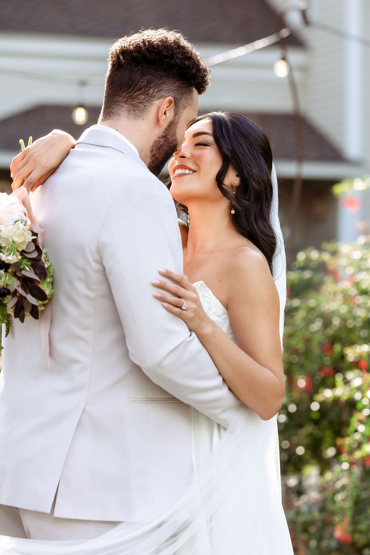 A tender embrace between newlyweds in white attire against a garden backdrop.