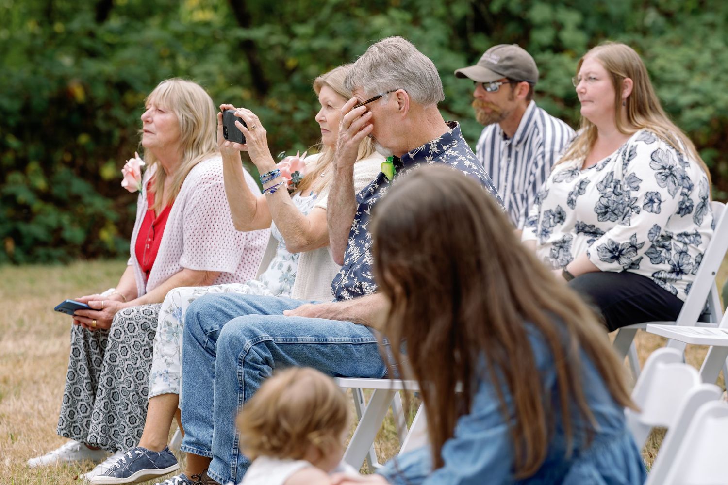 A group of people sit at tables outdoors enjoying a social gathering in a park-like setting.