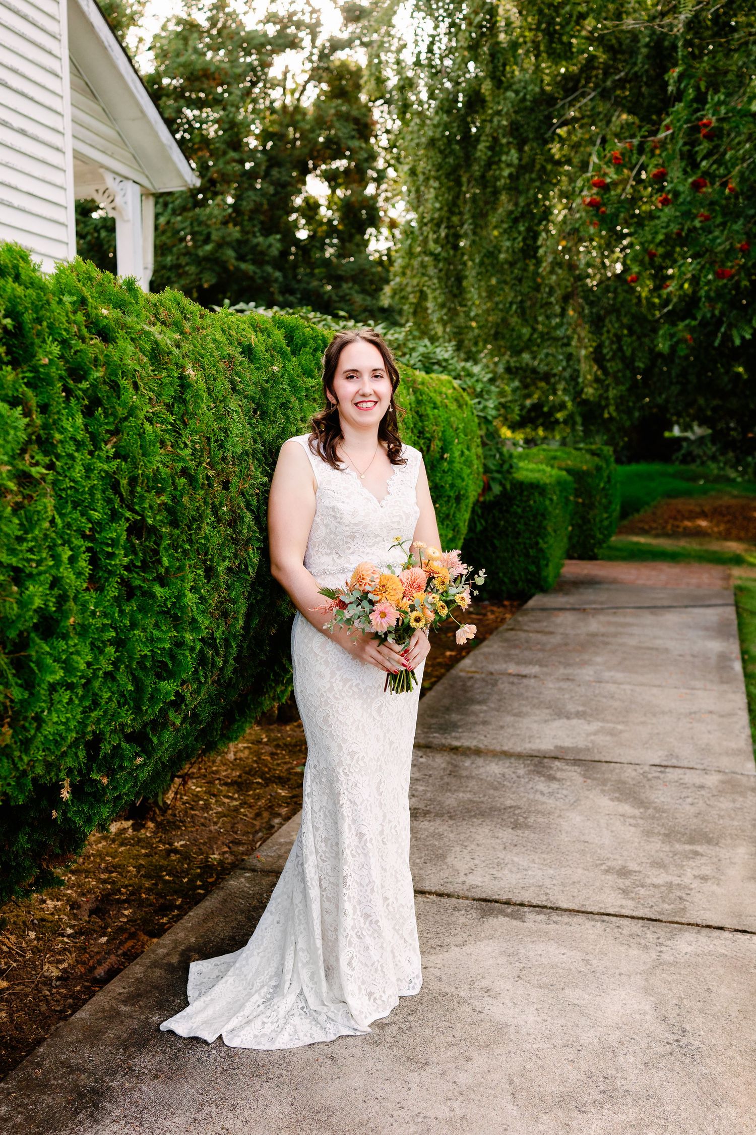 A bride in a white lace dress holds a peach and orange bouquet while standing on a garden path.