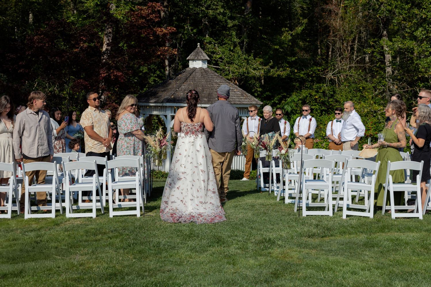 Outdoor wedding ceremony setup with white chairs and gazebo surrounded by trees.