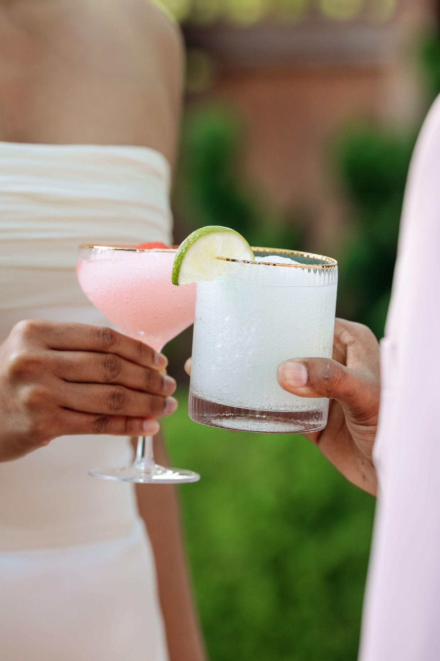 Hands holding elegant cocktail glasses with pink and clear drinks garnished with lime slices.