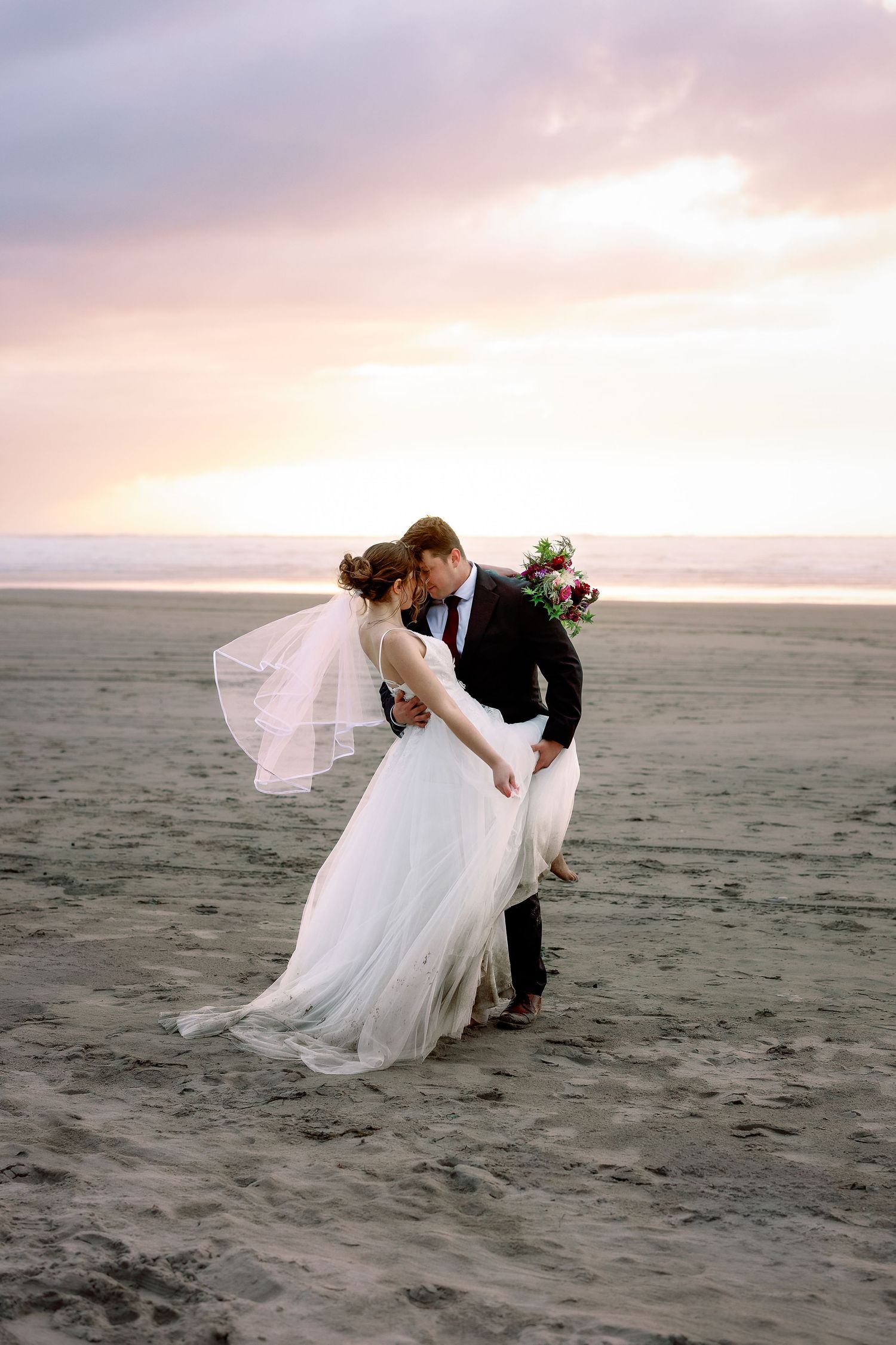 A romantic beach wedding photoshoot at sunset with a couple in formal attire posing on the sandy shore.