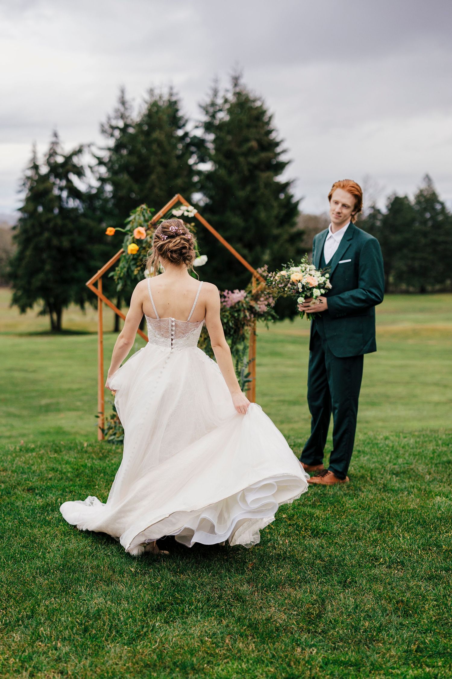 A bride in a flowing white gown twirls on a grassy lawn near a wooden arch with floral decorations.