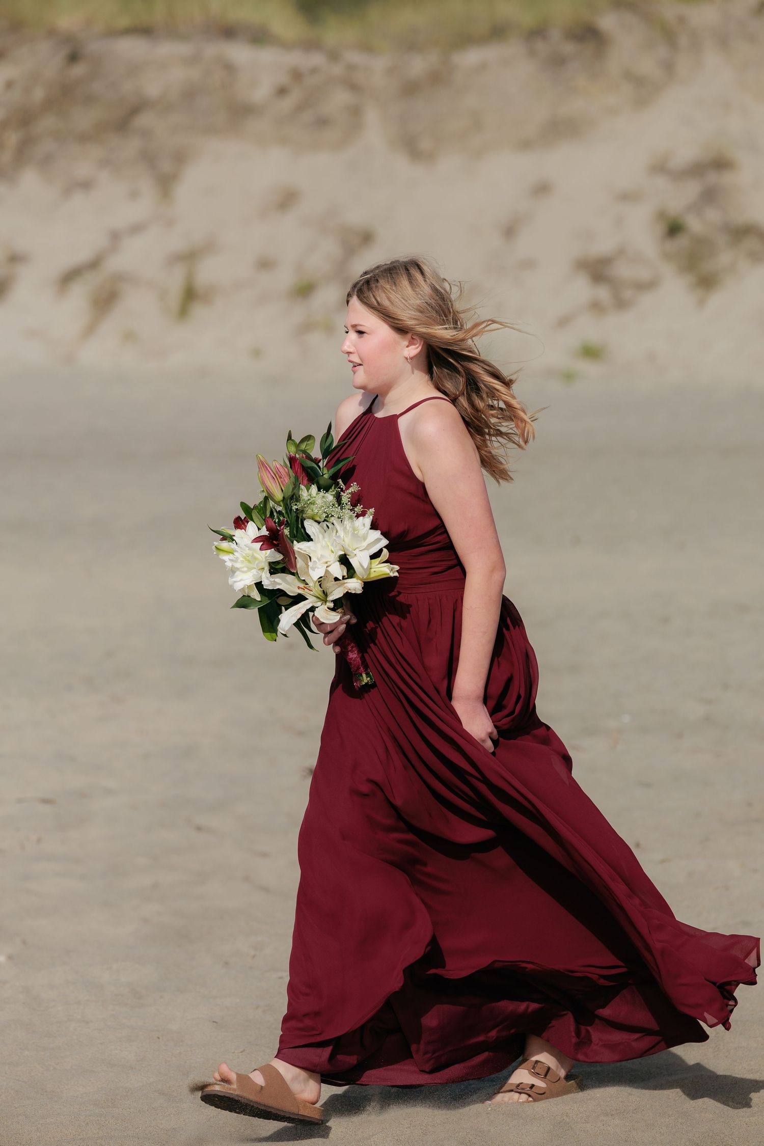 A person in a flowing burgundy dress walks on the beach holding a white floral bouquet.