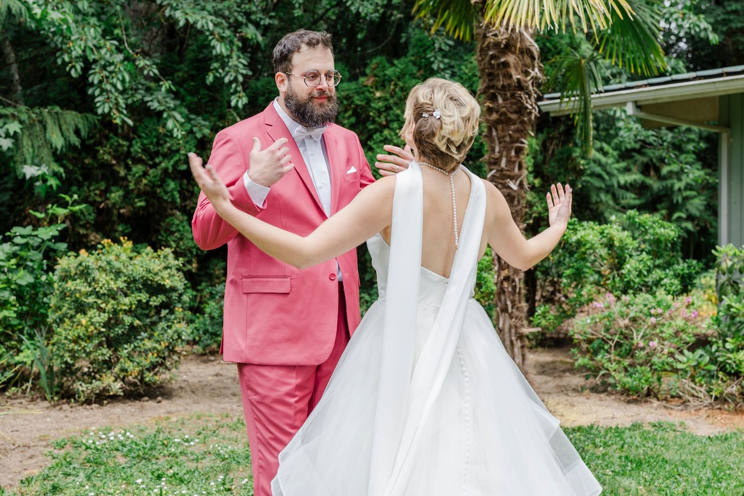 A couple dressed in a white gown and coral pink suit dance joyfully together in a lush garden setting.