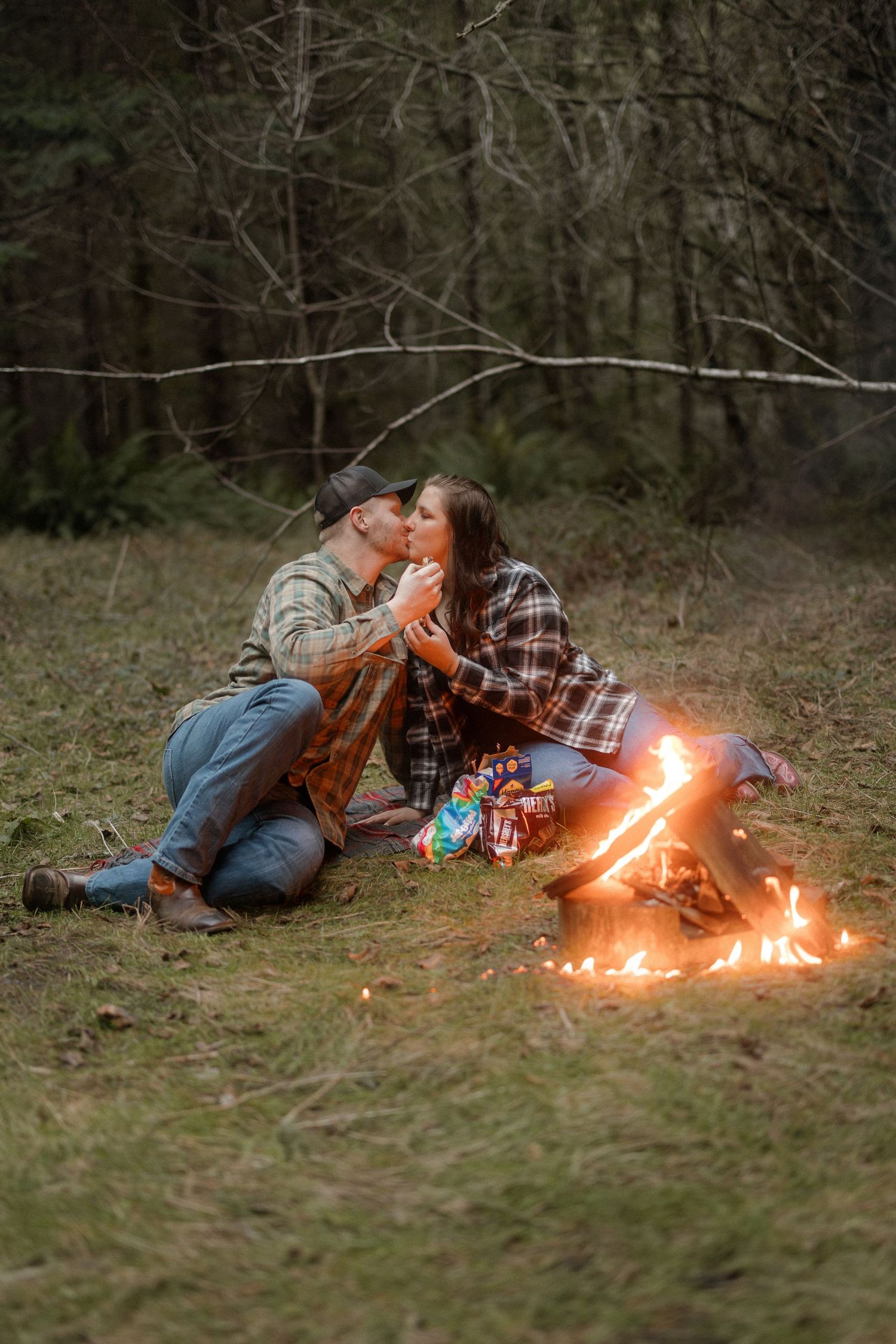 Couple enjoying a cozy evening by a campfire with drinks and plaid flannel in a wooded outdoor setting.