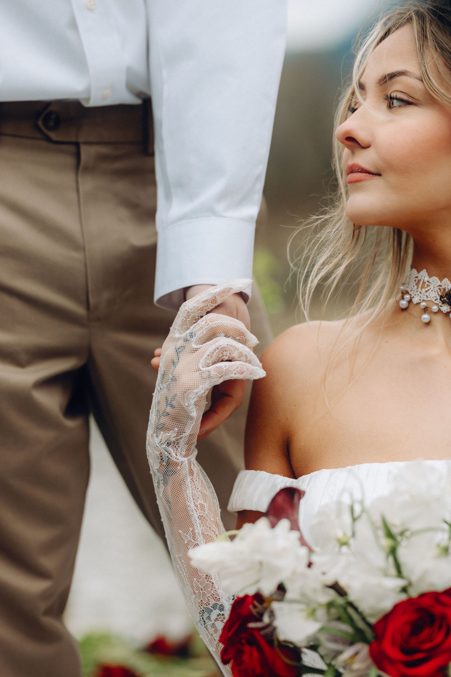 A wedding couple holds hands showing intricate lace sleeve details and red and white rose bouquet.