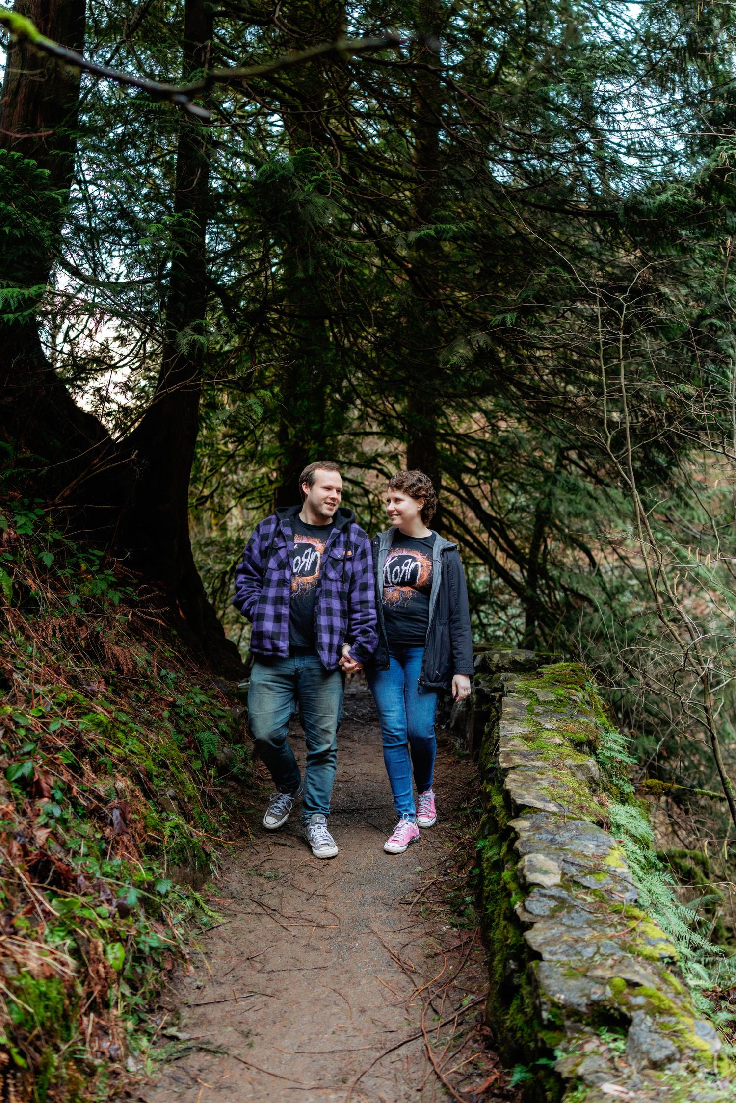 A couple walks along a scenic forest trail surrounded by lush greenery and mossy rocks.