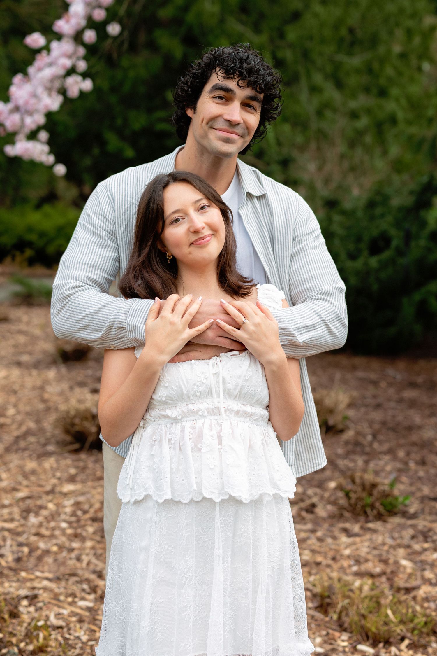 A romantic couple in white outfits embraces outdoors with pink cherry blossoms blooming in the background.