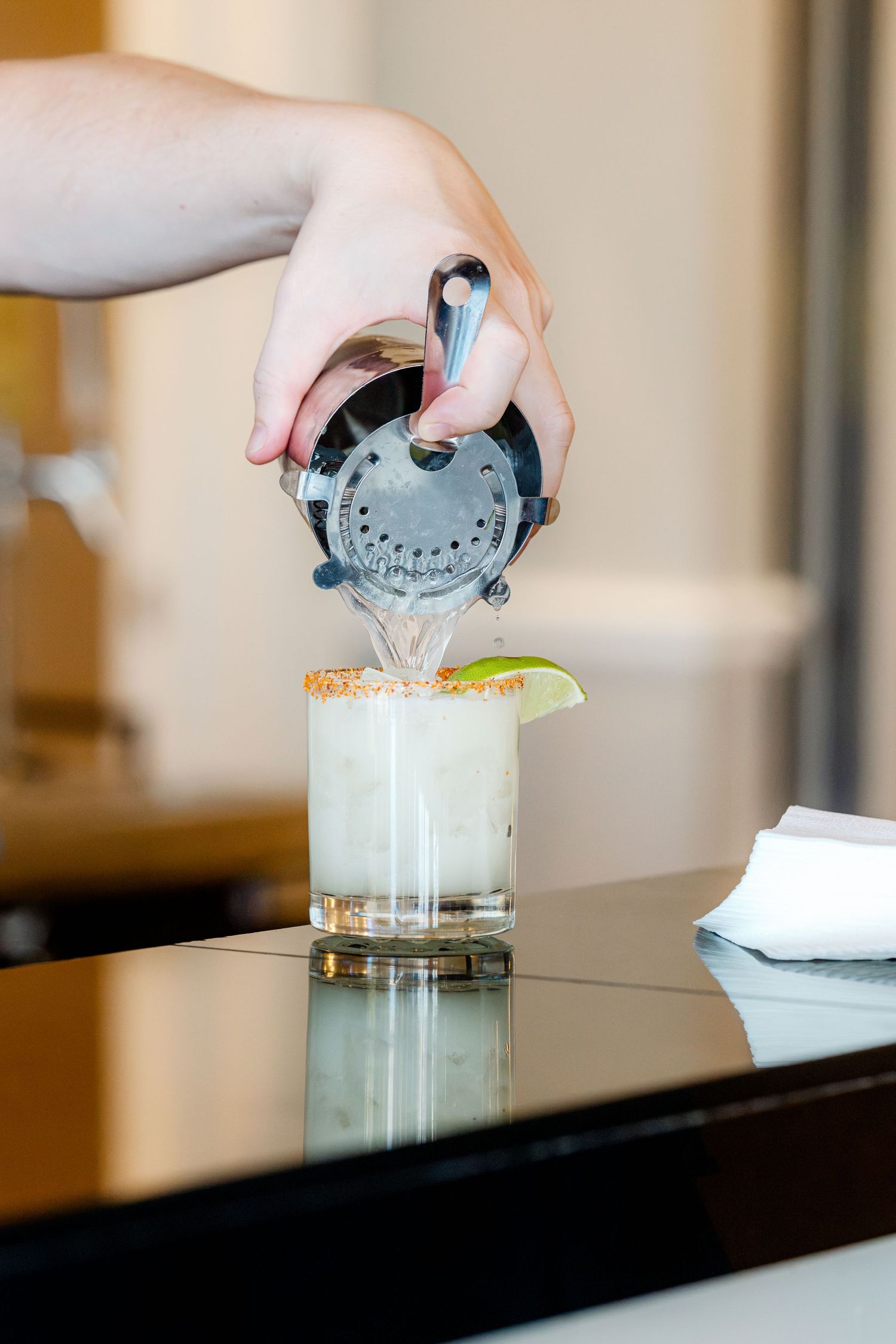 Bartender pouring a margarita cocktail with salted rim and lime garnish into a glass on a dark bar.