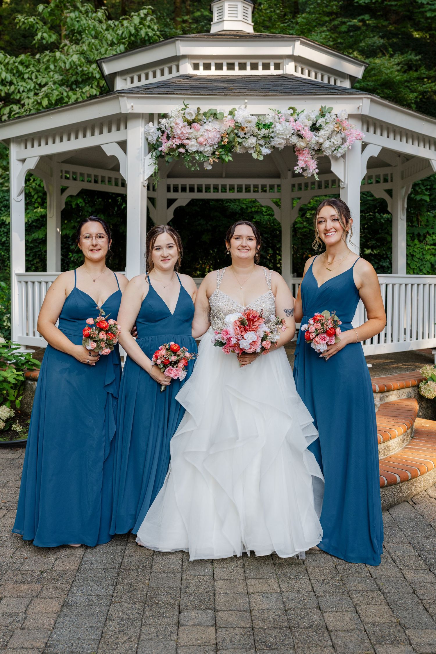 Bride in white gown with three bridesmaids in blue dresses holding pink bouquets before a floral gazebo.