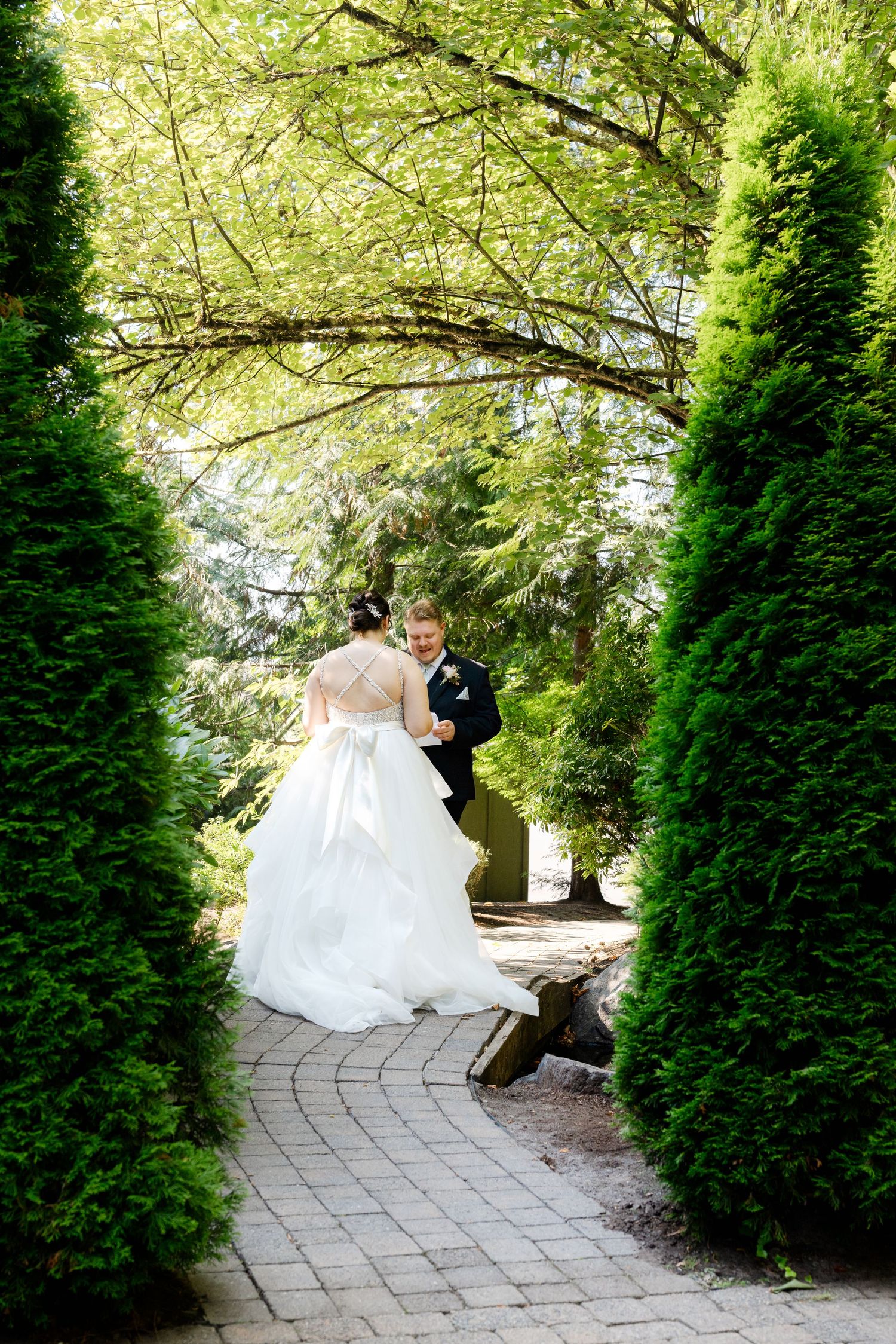 Bride and groom share a romantic moment on a brick path surrounded by lush green hedges.