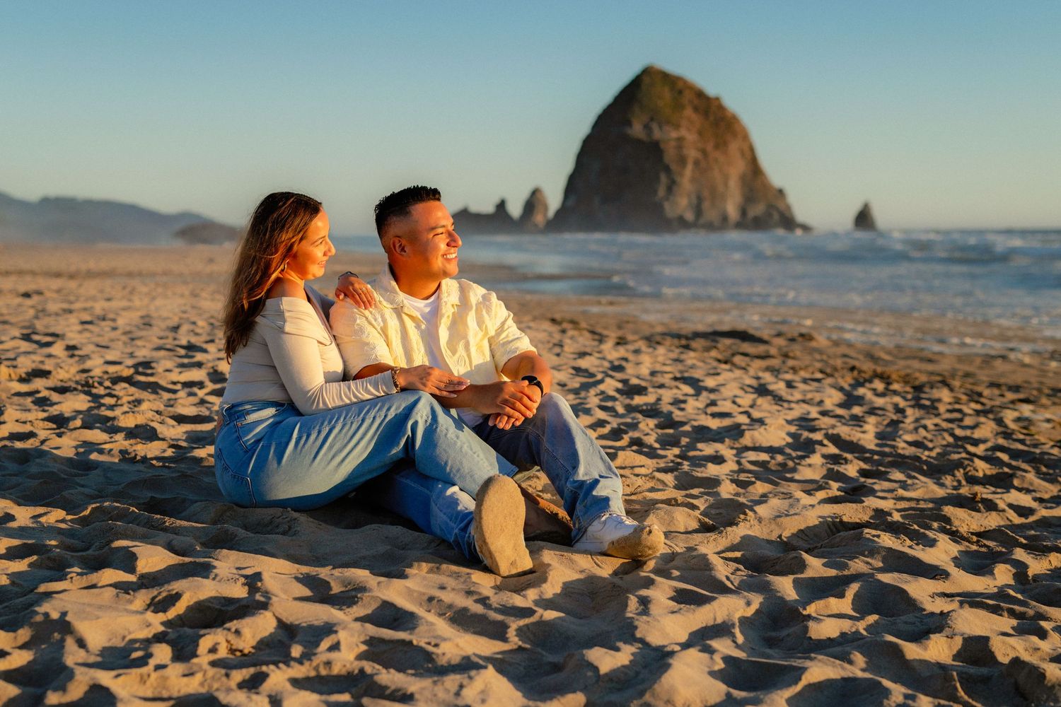 Engaged couple sitting together on Cannon Beach sand watching sunset near Haystack Rock.