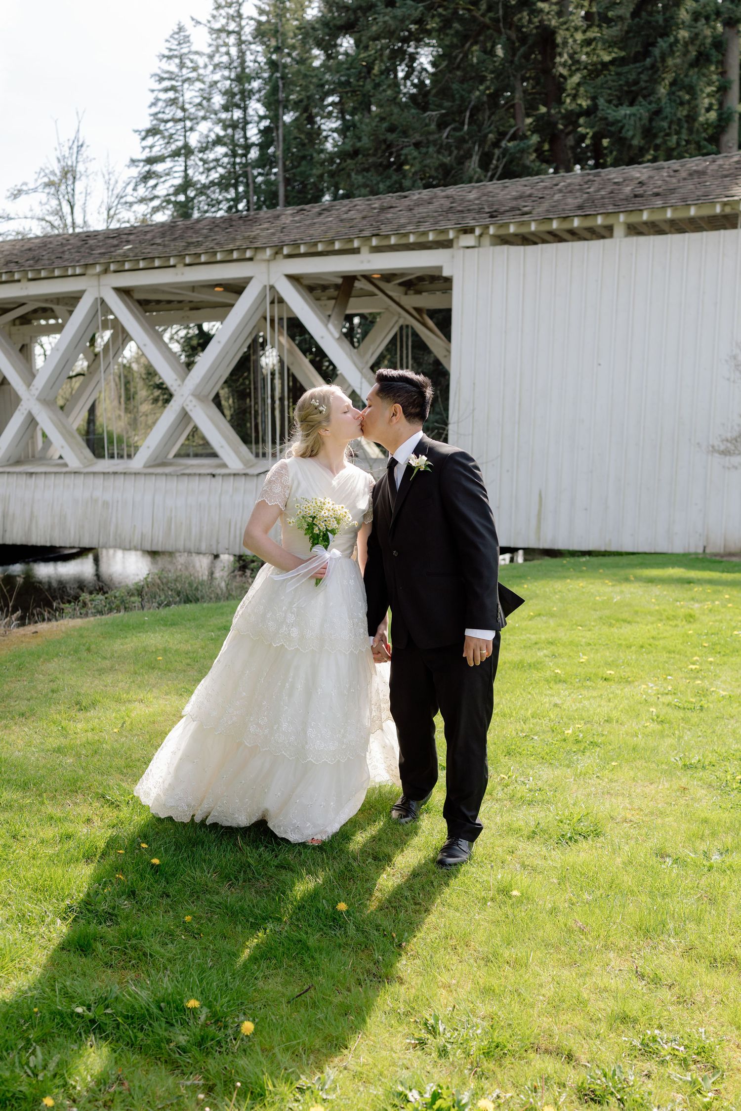 A bride and groom share a romantic kiss on a green lawn with a white covered bridge in the background.