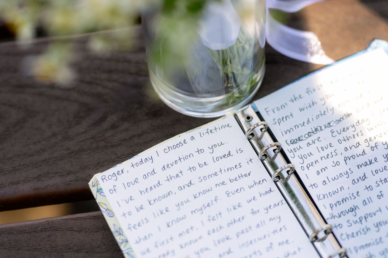 Open wedding vow notebook on wooden table with handwritten promises, beside a glass vase with white flowers.