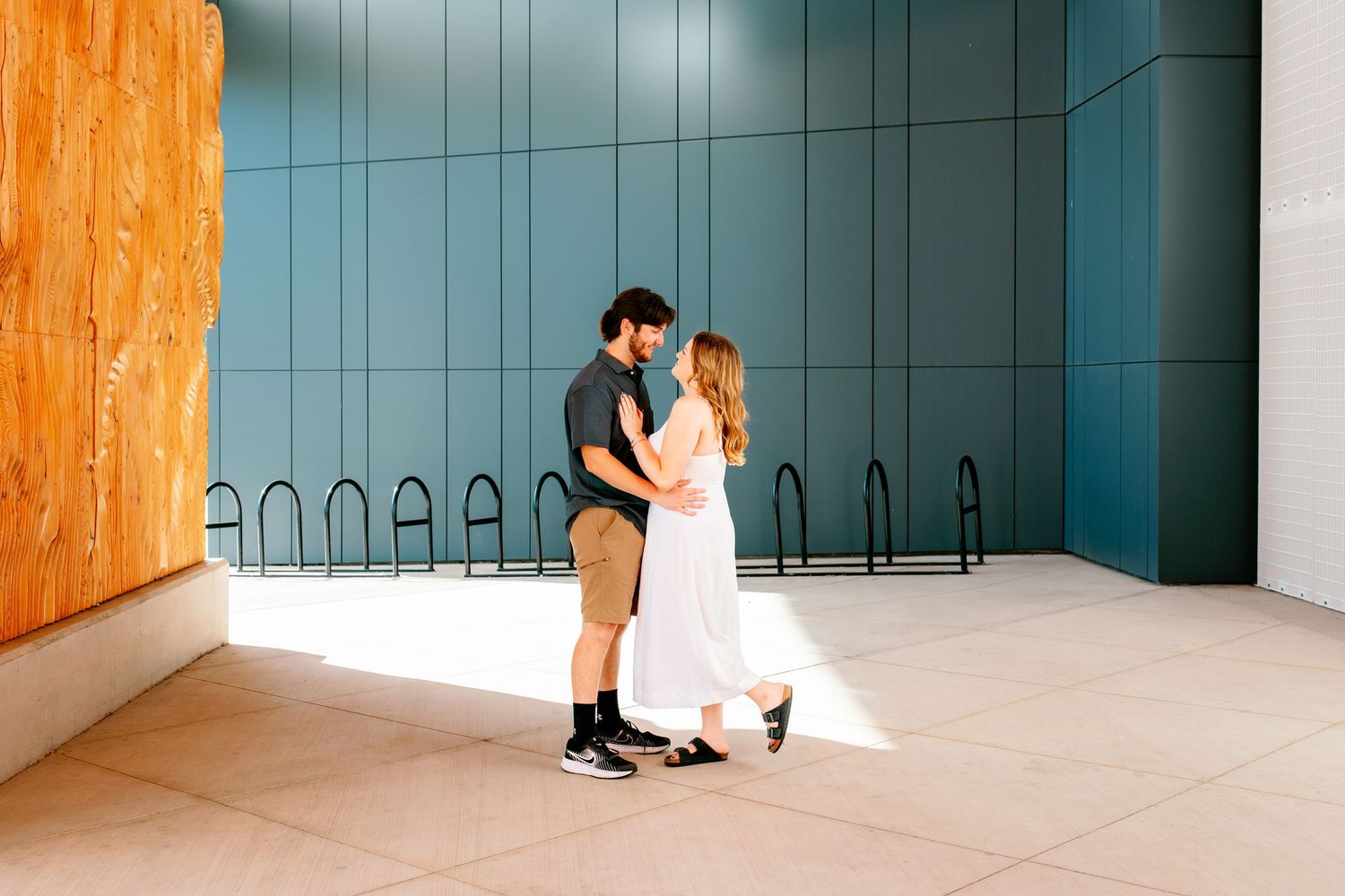 A couple embraces tenderly outside a modern building with teal panels and bike racks in warm sunlight.