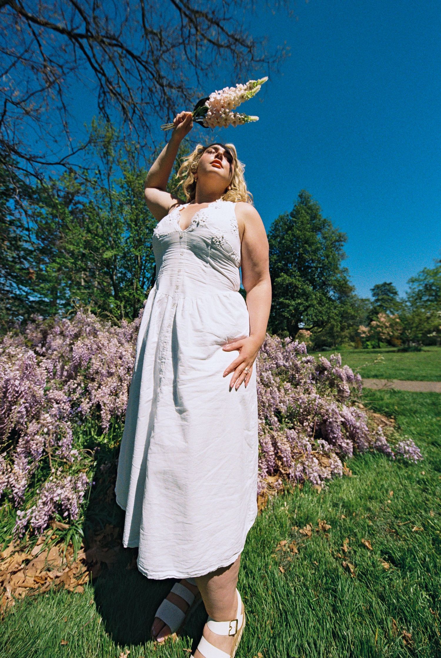 A woman in a white dress holds wildflowers among blooming purple wisteria under a blue sky.