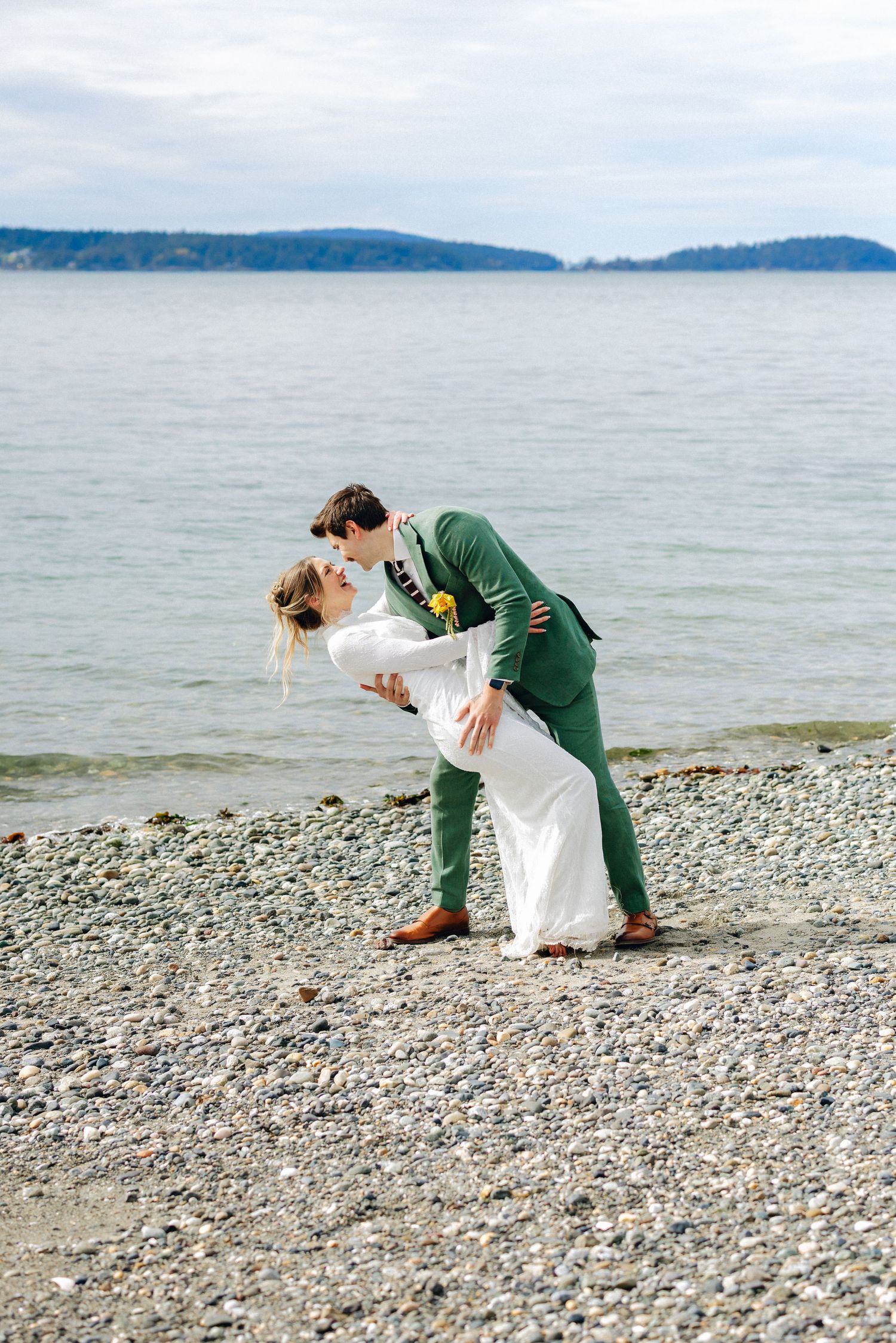 Bride and groom share romantic dip kiss on rocky beach shoreline with ocean view behind them.