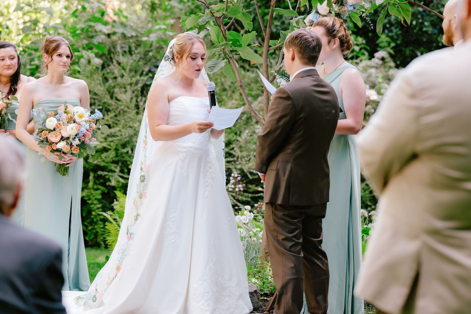 Bride reads vows to groom during outdoor garden wedding ceremony with bridesmaids in sage green dresses.
