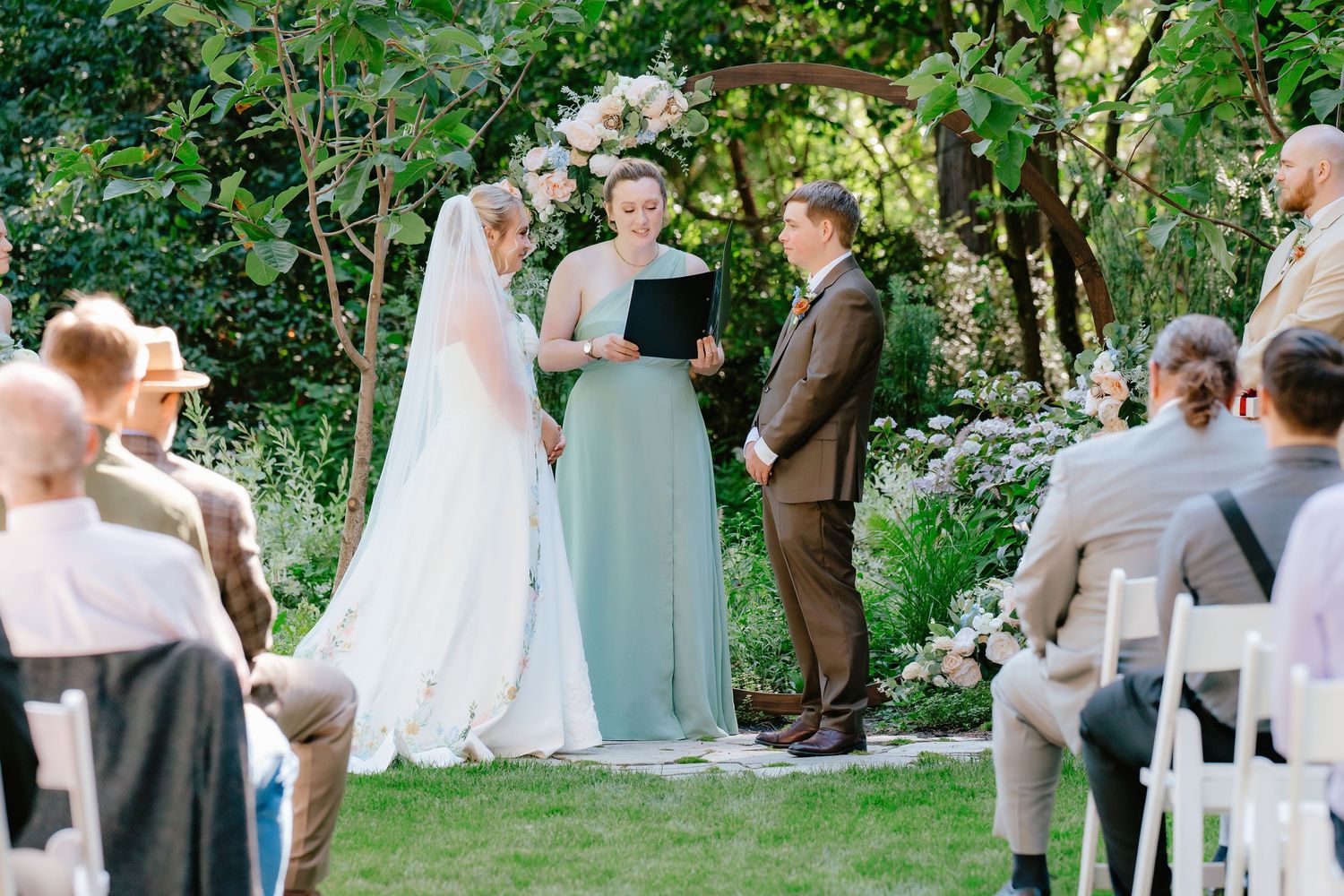 Outdoor garden wedding ceremony with bride in white gown, groom in brown suit, and officiant in mint dress under floral arch.