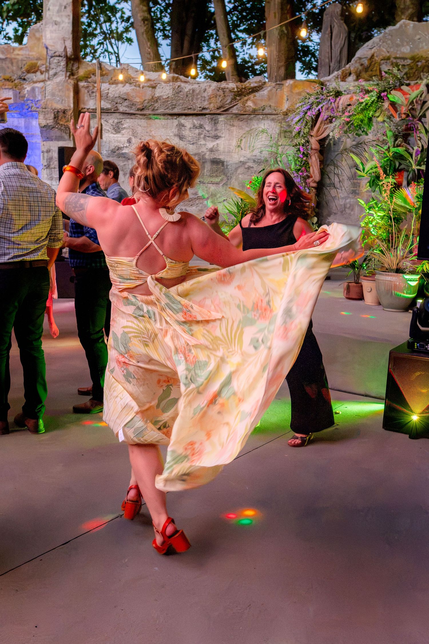 Women dancing joyfully at an outdoor evening party, one twirling a floral dress under string lights.