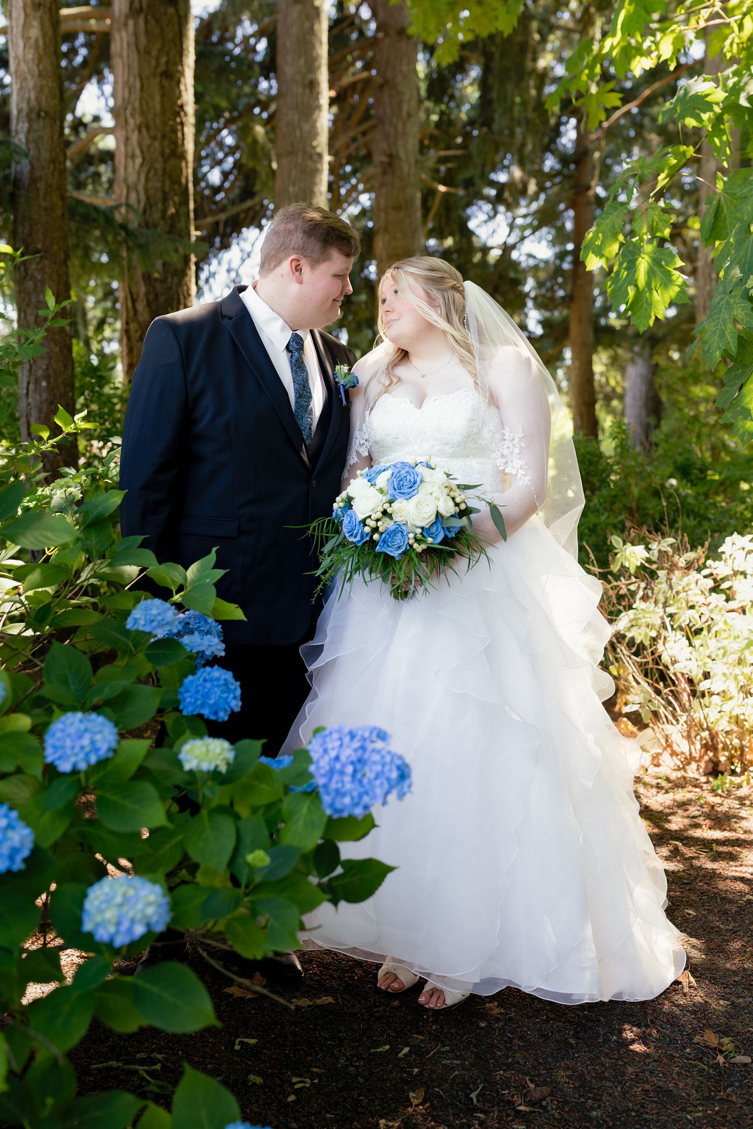 Bride and groom share a romantic moment in a garden with blue hydrangeas, bride holding a matching blue and white bouquet.