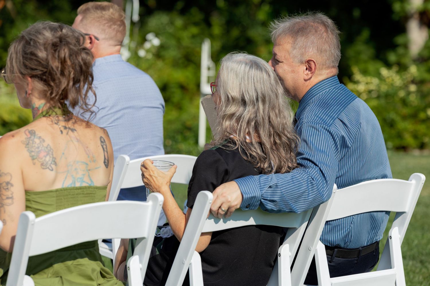 A couple sits close together on white chairs in an outdoor garden setting during a special event.