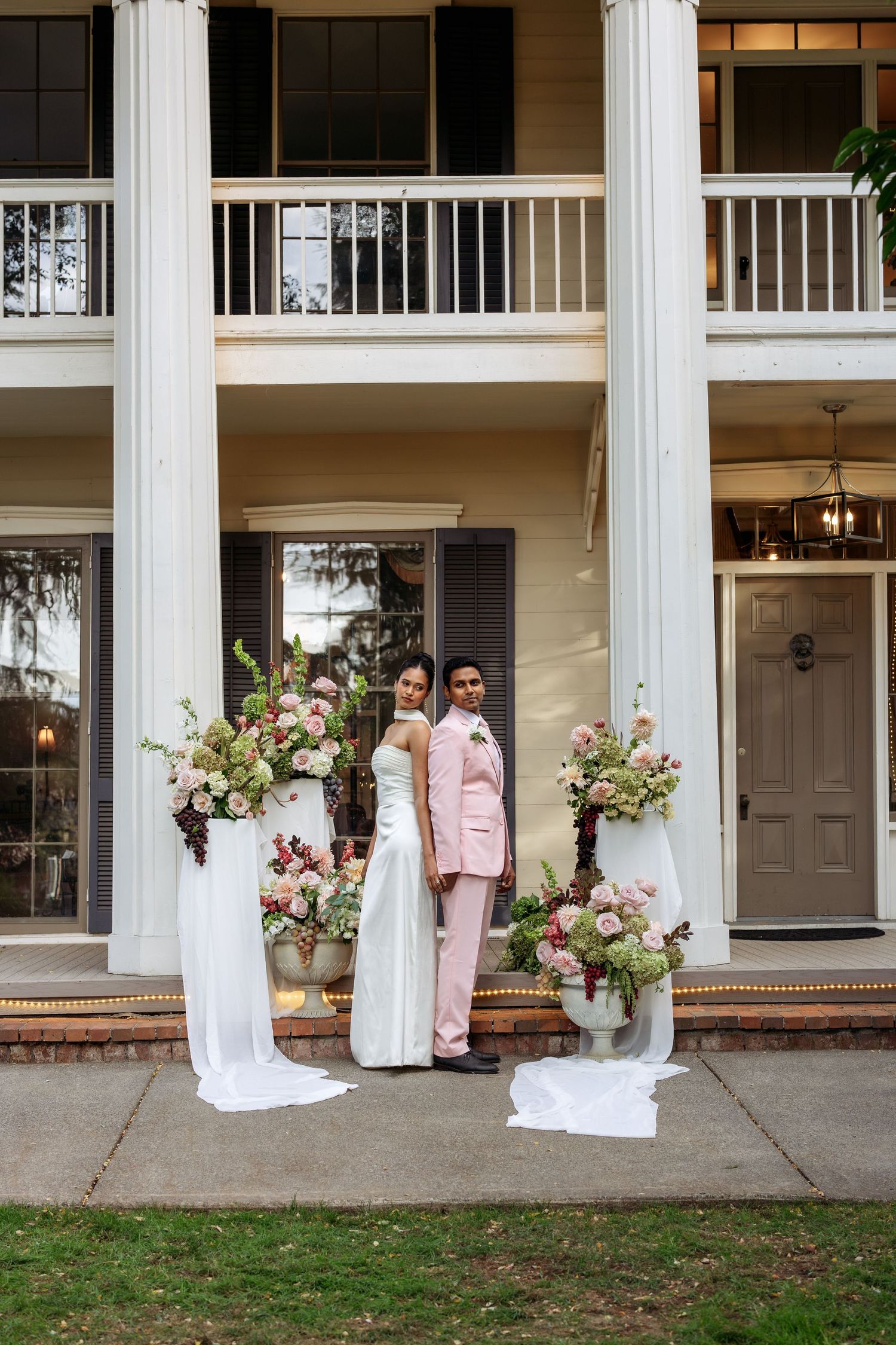 Wedding party poses on classic Southern mansion porch with white columns and floral arrangements.