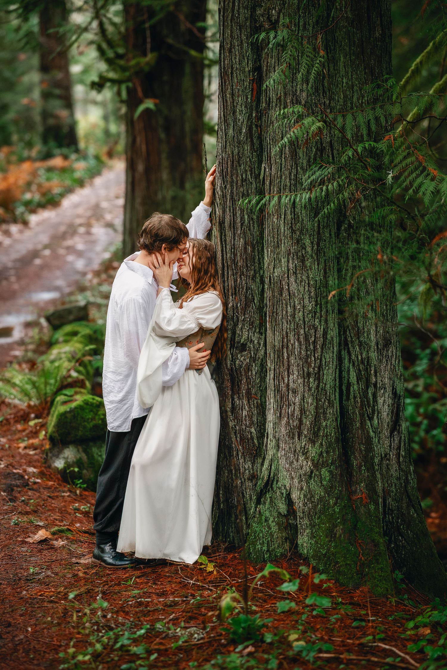 Two people in light clothing share an intimate moment against large tree trunks on a forest path.