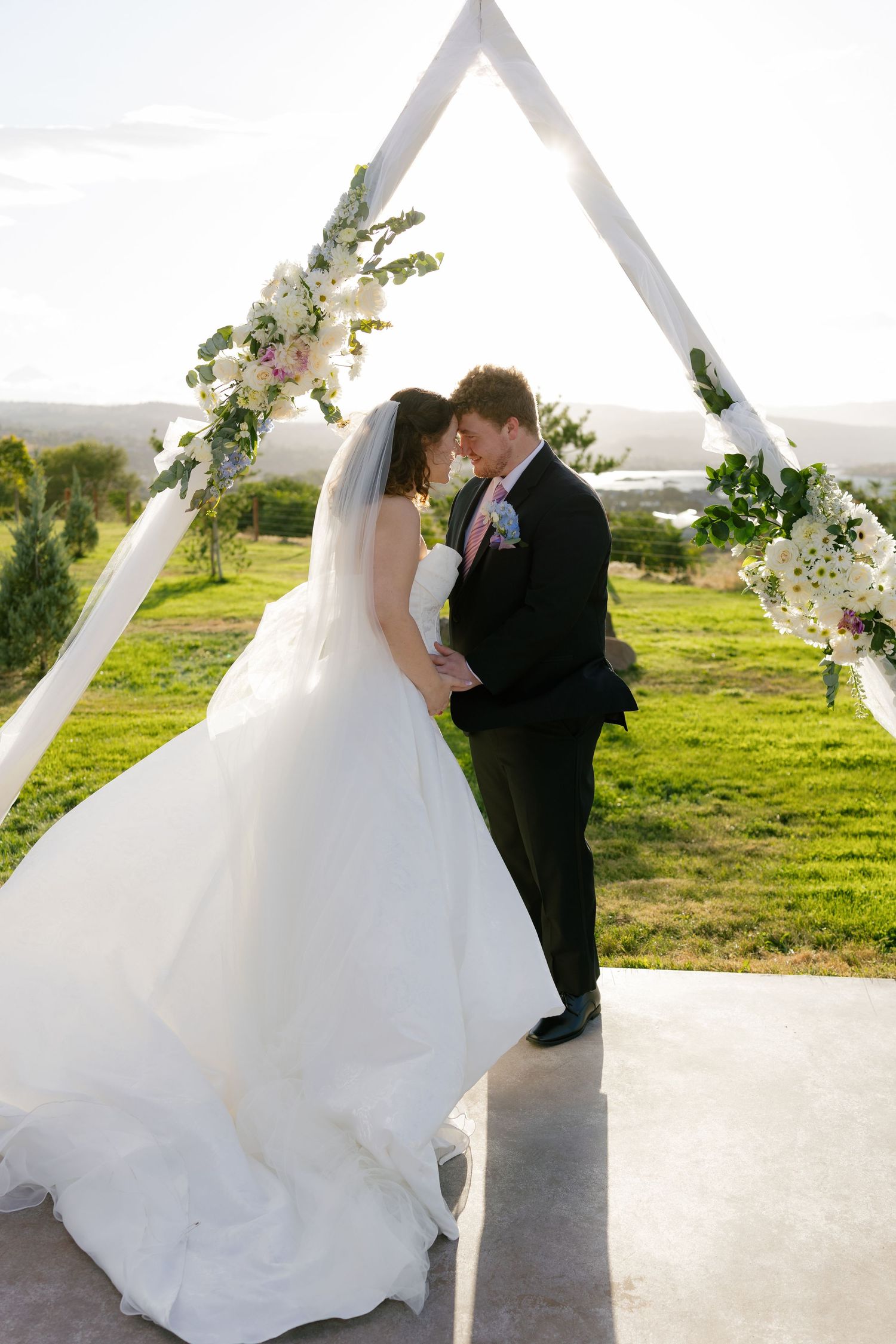 A couple shares a romantic moment during their outdoor wedding ceremony with flowing white dress and black suit.